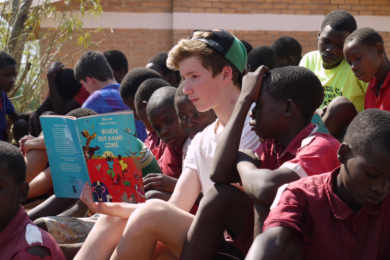 A guest sitting with a group of children outside reading a book out loud.