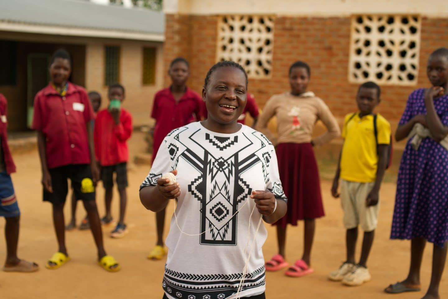Children are in a semicircle around the facilitator of the BRAVE programme who is smiling and explaining an activity to them. They are all standing outside infront of a school building.