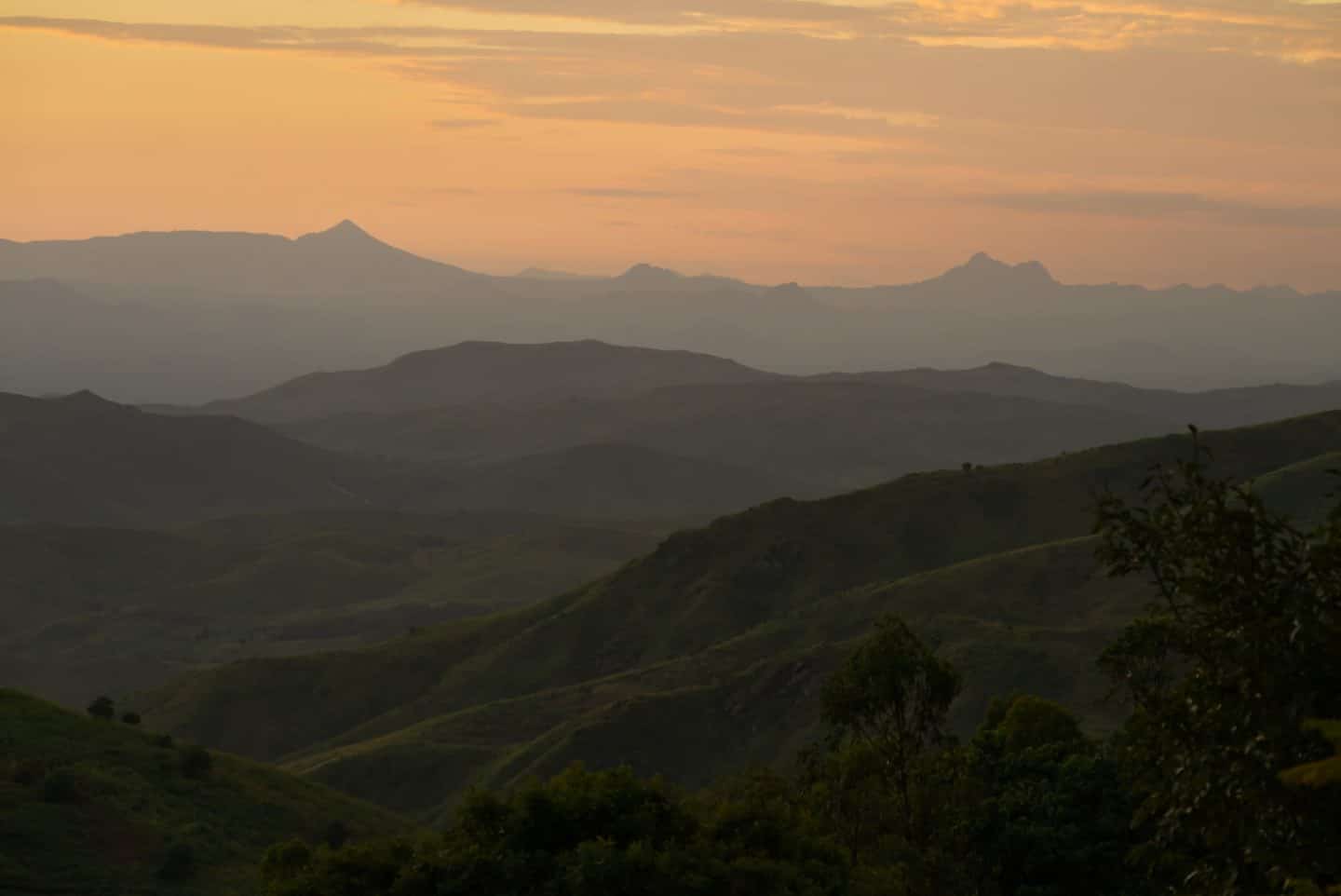 The view from Fisherman's Rest just after sunset, the sky is pink and orange with expansive views of mountains in the distance and the tops of hills in the foreground and middle distance lit with gentle taces of the fading sunlight. A place to travel with purpose.