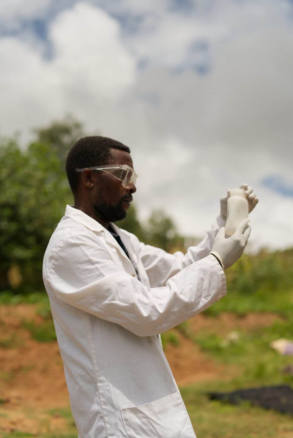 A Madzi Aplipo team member is wearing a lab coat holding up a sample of water in the outdoors ready to do water quality testing.
